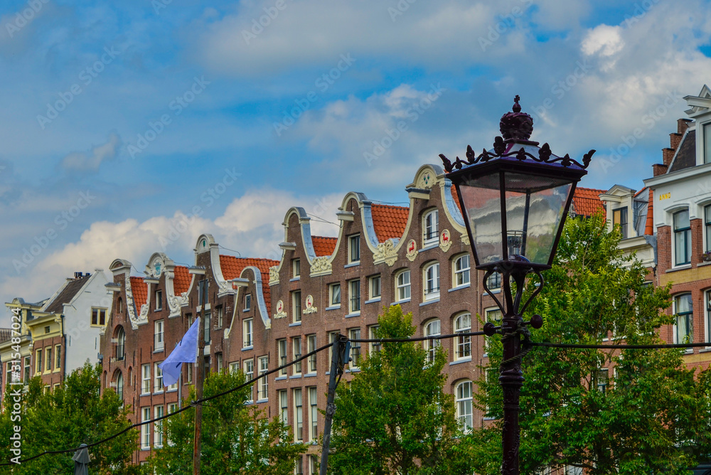 Amsterdam, Holland, August 2019. The typical and charming houses: they are a symbol of the city represented on a postcard. With brightly colored brick facades and distinctive roofs. A lamppost frames.