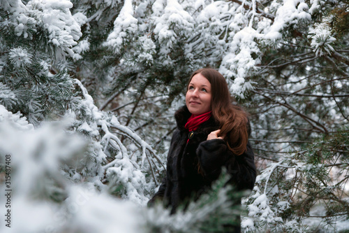 A beautiful woman standing among snow-covered trees in a winter forest and enjoying the first snow. Model with long hair. A girl in a fur coat. Wanderlust.