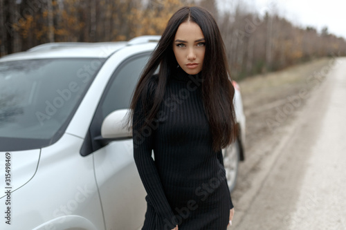 A beautiful girl with long dark hair is standing by a white car