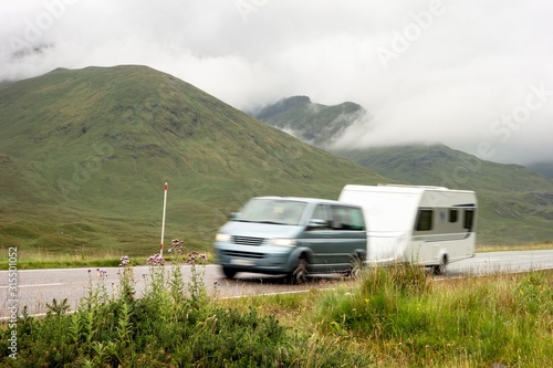 A blue MPV car with a trailer caravan driving on a road in the landscape of Scottish Highlands in misty weather with motion blur effect