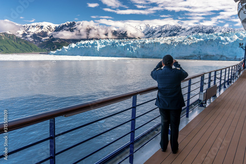 Woman is wearing a stripes and taking a picture of Hubbard Glacier on her mobile phone. Lady is crew member on a cruise ship. Working as a ships officer. Snowy mountains and iceberg.