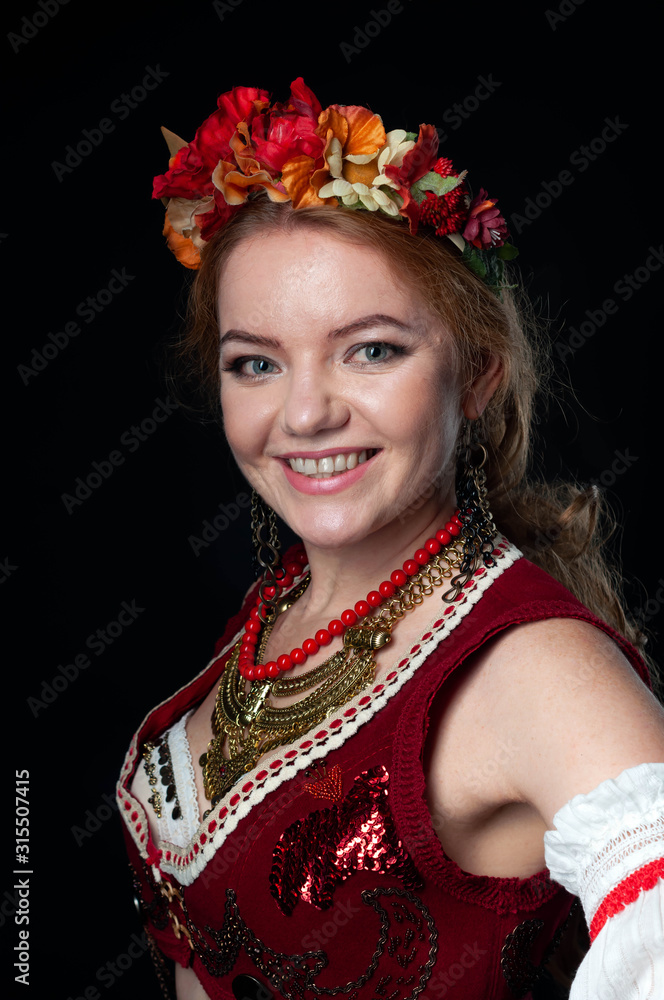 Naklejka premium portrait of attractive smiling woman in traditional costume in ukranian national wreath with flowers, red vest and golden necklace looking at camera isolated on black