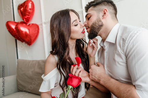 man proposes to woman with ring and red rose at home on couch with heart shaped balloons
