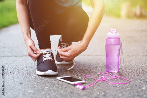 runner woman tying running shoes laces getting ready for race on asphalt with bottle of water and phone with headphones