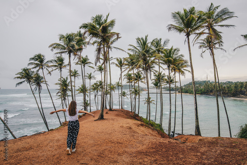 Back view of young yourist woman visiting Coconut tree hill in Mirissa, Sri Lanka. Coconut Tree Hill Mirissa is a unique cliff located at the end of a beach which is covered in palm trees. photo