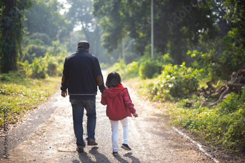 Back side of an Indian Bengali father and his little daughter with winter garments are walking through a pathway within a green natural forest in a winter morning. Indian lifestyle and winter.