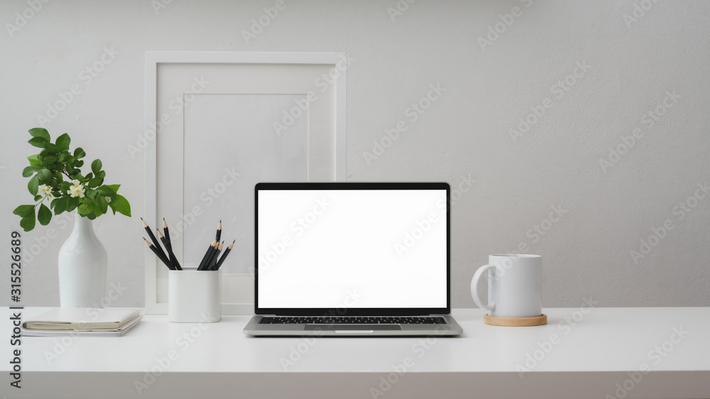 Cropped shot of workspace with blank screen laptop, frame, pencils, coffee cup and vase on white table with white wall Cropped shot of workspace with blank screen laptop, frame, pencils, coffee cup and vase on white table with white wall