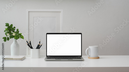 Cropped shot of workspace with blank screen laptop, frame, pencils, coffee cup and vase on white table with white wall © bongkarn
