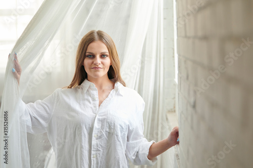 Stunning young Caucasian woman poses in studio wearing a men's white button-up shirt - near window