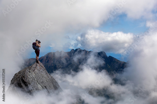 Adventure Female Photographer and Hiker standing on top of a high rocky mountain peak during a vibrant sunny summer day. Composite Image. Photos taken from British Columbia, Canada.