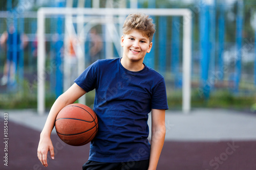 Cute boy in blue t shirt plays basketball on city playground. Active teen enjoying outdoor game with orange ball. Hobby, active lifestyle, sport for kids.  © Natali