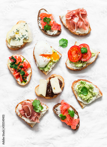 Variety of tapas sandwiches - sandwiches with prosciutto, avocado, salmon, egg, tomatoes, jamon, gorgonzola, brie, pear on a light background, top view. Delicious snack, appetizers