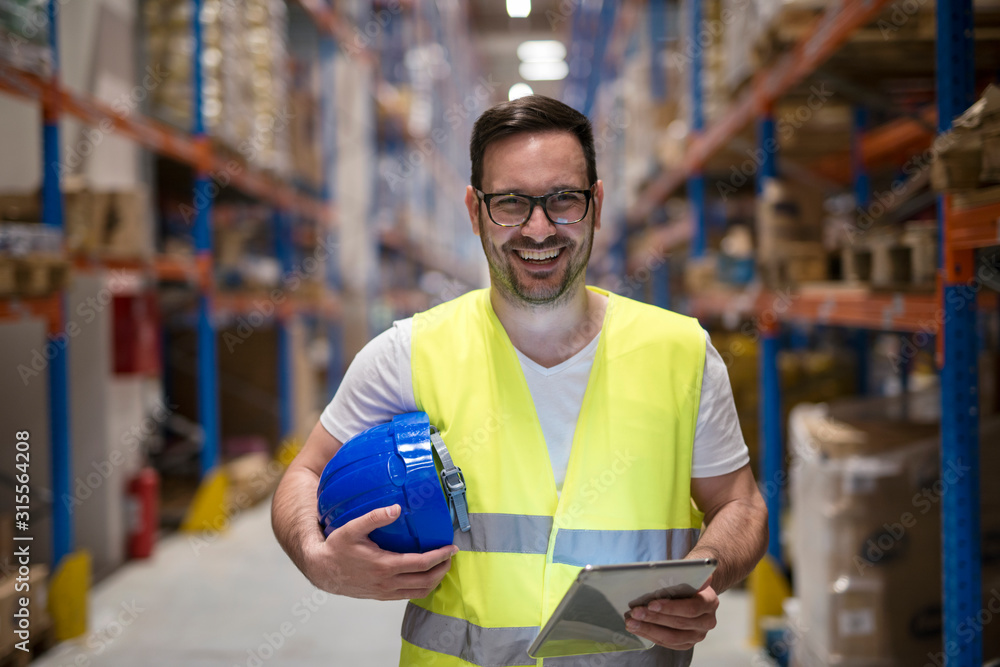 Portrait of smiling caucasian warehouse worker with tablet standing in storage department. - obrazy, fototapety, plakaty 