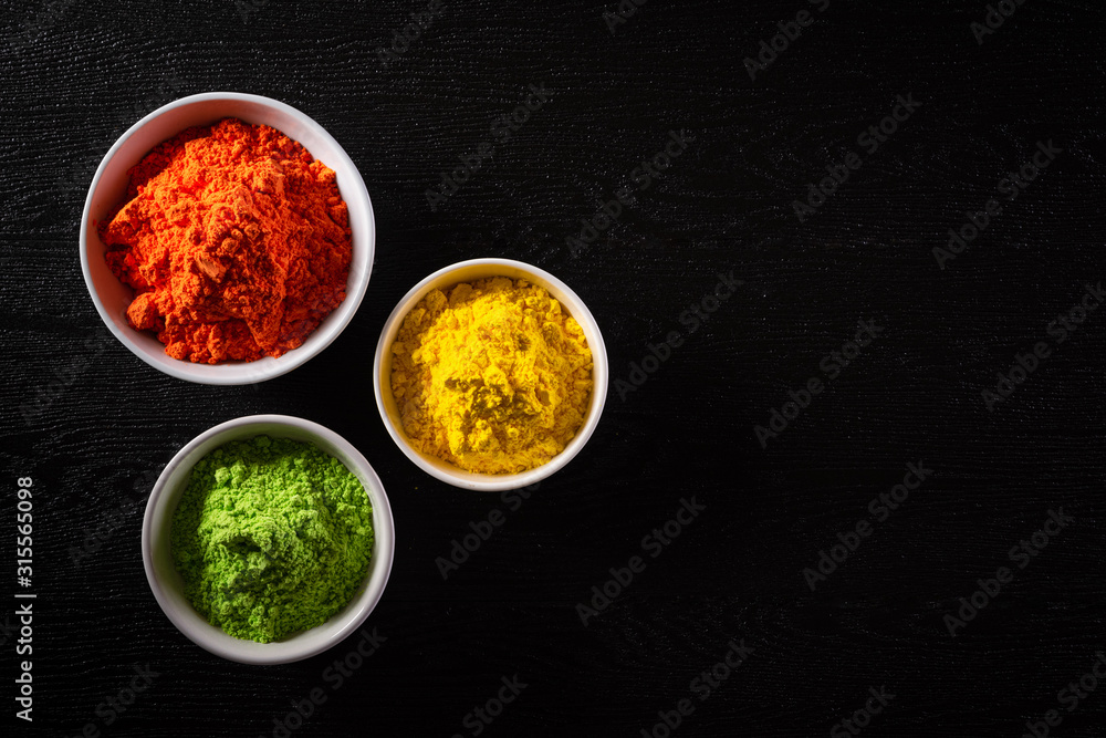Holi festival celebration. Traditional Indian Holi colours powder decoration with paints. Top view of Organic Gulal colors in bowls, spices, rustic on black background for Holi festival. Holi festival celebration. Traditional Indian Holi colours powder decoration with paints. Top view of Organic Gulal colors in bowls, spices, rustic on black background for Holi festival.