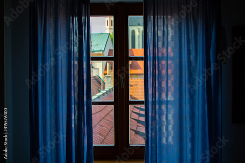 Curtained window from inside of a house. Roofs of houses through it