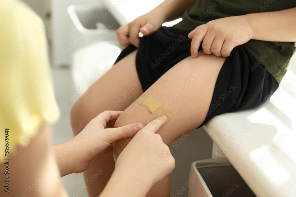 Fototapeta premium Sister putting sticking plaster onto little brother's leg indoors, closeup