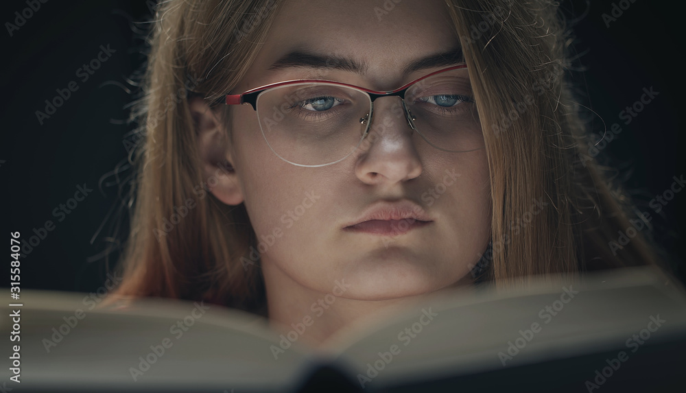 Close-up of concentrated girl in glasses behind book isolated on black background Close-up of concentrated girl in glasses behind book isolated on black background