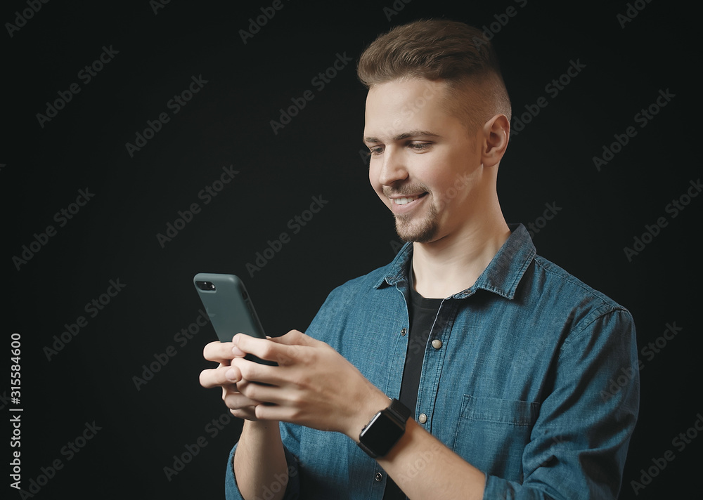 Smiling joyful guy in jeans shirt typing sms on smartphone, isolated black background Smiling joyful guy in jeans shirt typing sms on smartphone, isolated black background