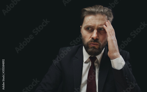 Portrait of thoughtful questioned bearded businessman in black suit touching his forehead against dark background