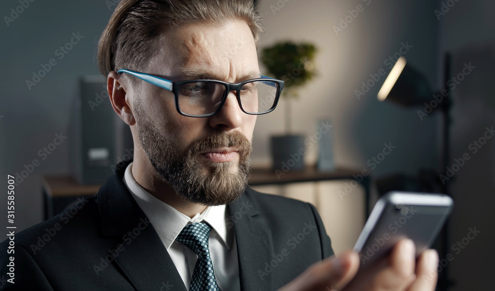 Serious bearded businessman in glasses being in office and looking at smartphone, wireless technology concept Serious bearded businessman in glasses being in office and looking at smartphone, wireless technology concept