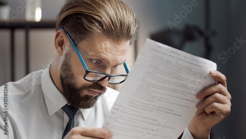 Close-up of astonished bearded businessman wearing glasses reading document with wide open eyes at work photo