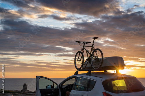 Mounted mountain bicycle silhouette on the car roof with evening sun light rays background. Safe sport items transportation using a car concept image.
