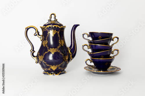 Closeup of a beautiful cobalt blue colored vintage porcelain tea set with golden floral pattern on white background. The set includes a tea pot, and a stack of tea cups.