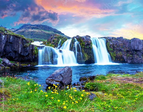 Beautiful terrific landscape with yellow flowers and big stone near waterfall Kirkjufell in Iceland at sunset. Exotic countries. Amazing places. Popular tourist atraction. 