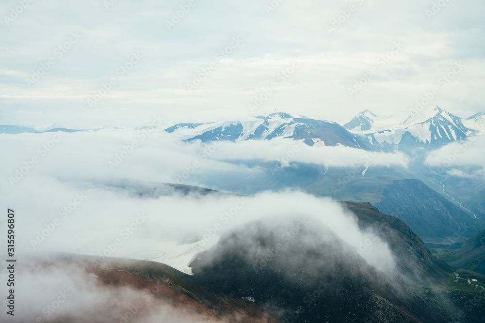 Obraz premium Beautiful atmospheric alpine landscape to big snowy mountain ridge with glacier. Giant low clouds among rockies in valley. Wonderful tranquil highland scenery. Flying over mountains above clouds.