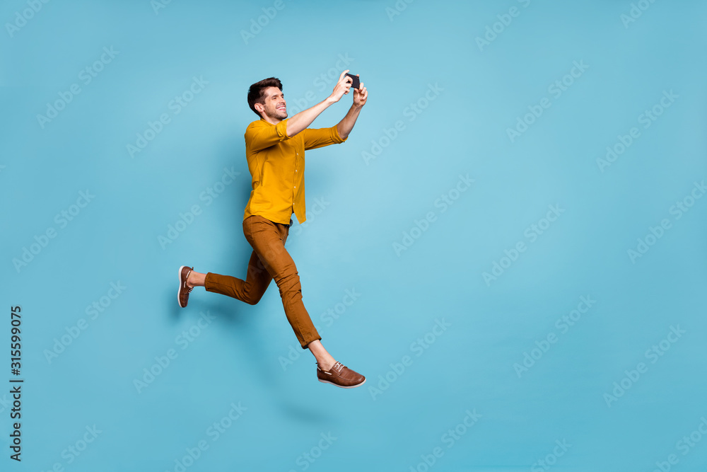 Full size profile photo of funny guy jumping high holding telephone making selfies online multimedia translation wear yellow shirt pants isolated blue color background