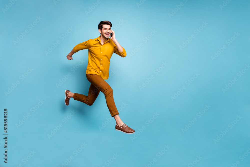 Fototapeta premium I'm on my way. Full length photo of crazy guy jumping high rushing to party holding telephone speaking friends wear yellow shirt pants isolated blue color background
