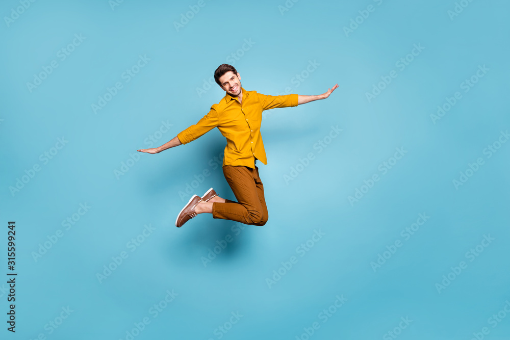 Full length photo of crazy guy jumping high holding hands spread by sides pretending bird flight wear yellow shirt trousers isolated blue color background Full length photo of crazy guy jumping high holding hands spread by sides pretending bird flight wear yellow shirt trousers isolated blue color background