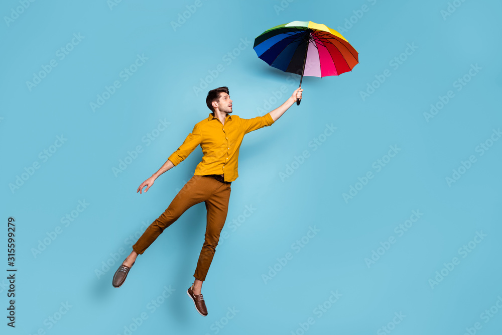 Full length photo of crazy guy jumping high holding colored bright umbrella flying up higher wear yellow shirt trousers isolated blue color background Full length photo of crazy guy jumping high holding colored bright umbrella flying up higher wear yellow shirt trousers isolated blue color background