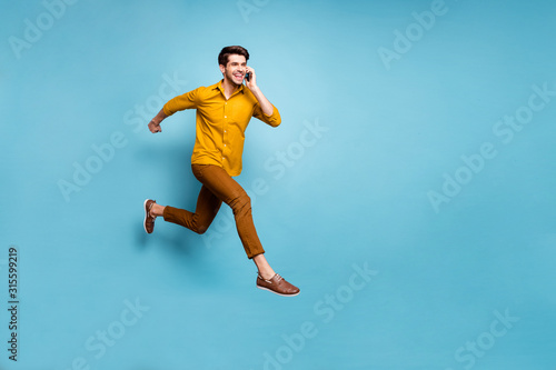 I'm on my way. Full length photo of crazy guy jumping high rushing to party holding telephone speaking friends wear yellow shirt pants isolated blue color background