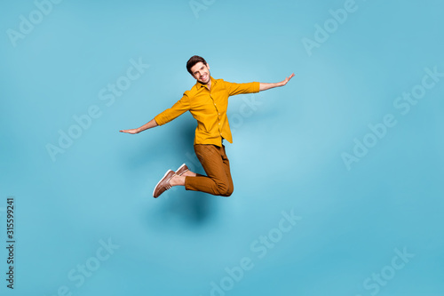 Full length photo of crazy guy jumping high holding hands spread by sides pretending bird flight wear yellow shirt trousers isolated blue color background