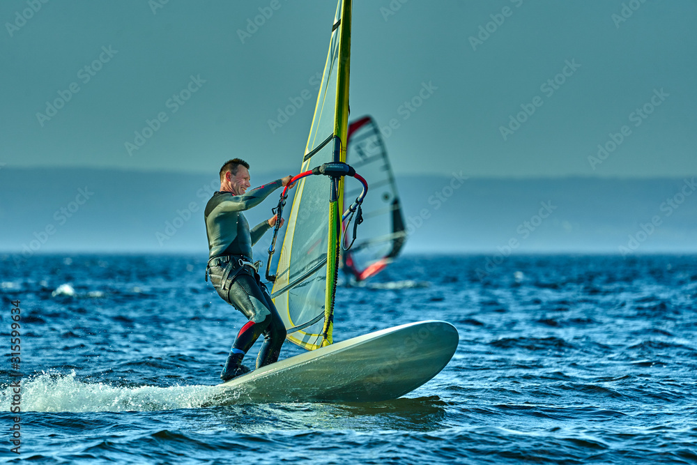 Naklejka premium A male athlete is interested in windsurfing. He moves on a Sailboard on a large lake on an autumn day.