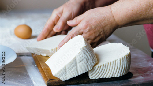 Close-up hands of senior female is cutting a white soft cheese for bakery preparation at domestic kitchen