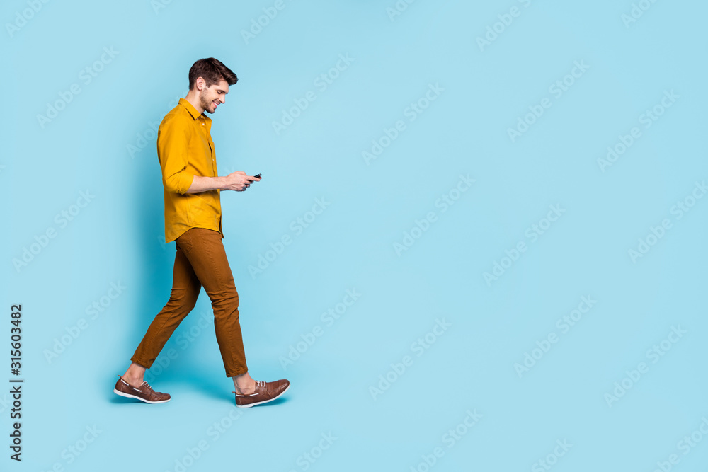 Photo of smiling toothy man wearing pants trousers footwear holding telephone with hands staring into screen walking into empty space isolated over blue pastel color background Photo of smiling toothy man wearing pants trousers footwear holding telephone with hands staring into screen walking into empty space isolated over blue pastel color background