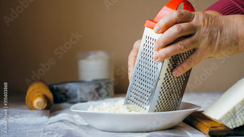 Close-up hands of mature woman is rubs a white soft cheese on a steel grater for preparing bakery at home kitchen
