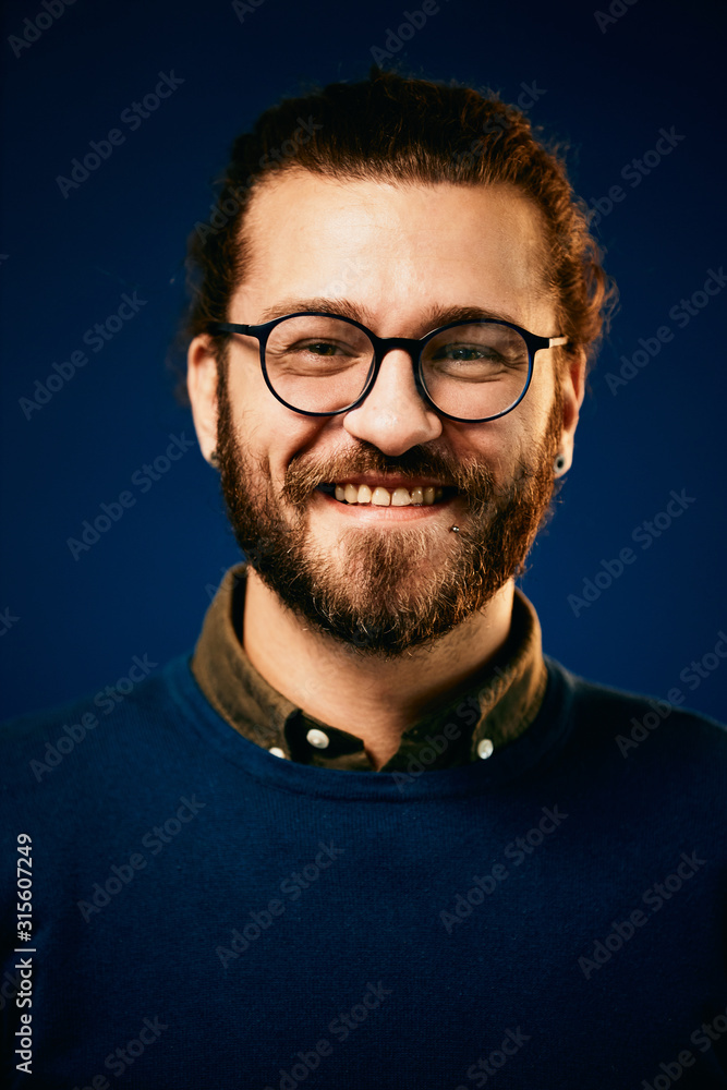 Obraz premium Portret całkiem uśmiechnięty brodaty hipster przed niebieskim tle i patrząc na kamery.