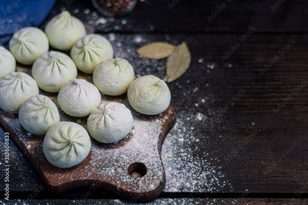 Frozen khinkali on a wooden board, sprinkled with flour. In the background pepper and bay leaf. On a black wooden background.