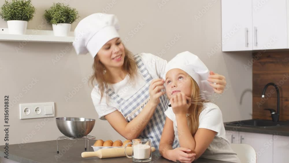 Beautiful woman in apron fixing cap of daughter preparing for baking cake, eggs and cookware on table Beautiful woman in apron fixing cap of daughter preparing for baking cake, eggs and cookware on table