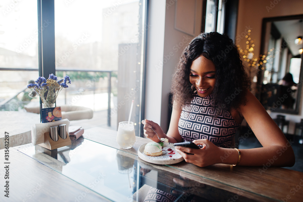 Сharming elegant young african american woman with long curly hair wearing jumpsuit sitting at cafe indoor eating dessert and look at mobile phone. Сharming elegant young african american woman with long curly hair wearing jumpsuit sitting at cafe indoor eating dessert and look at mobile phone.