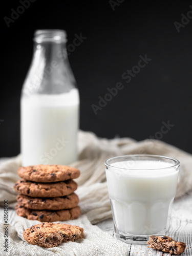 A glass with milk and several oatmeal cookies with chocolate on a wooden surface. Close-up. Bottle of milk in the background. Natural dairy products. Vertical orientation. Shallow depth of field.