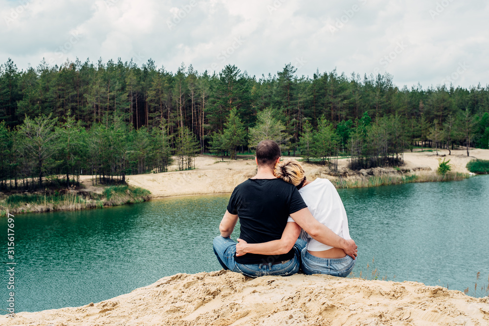 Girl and guy holding hands and sit on the shore of a forest lake. beautiful view of the lake . back view. Girl and guy holding hands and sit on the shore of a forest lake. beautiful view of the lake . back view.