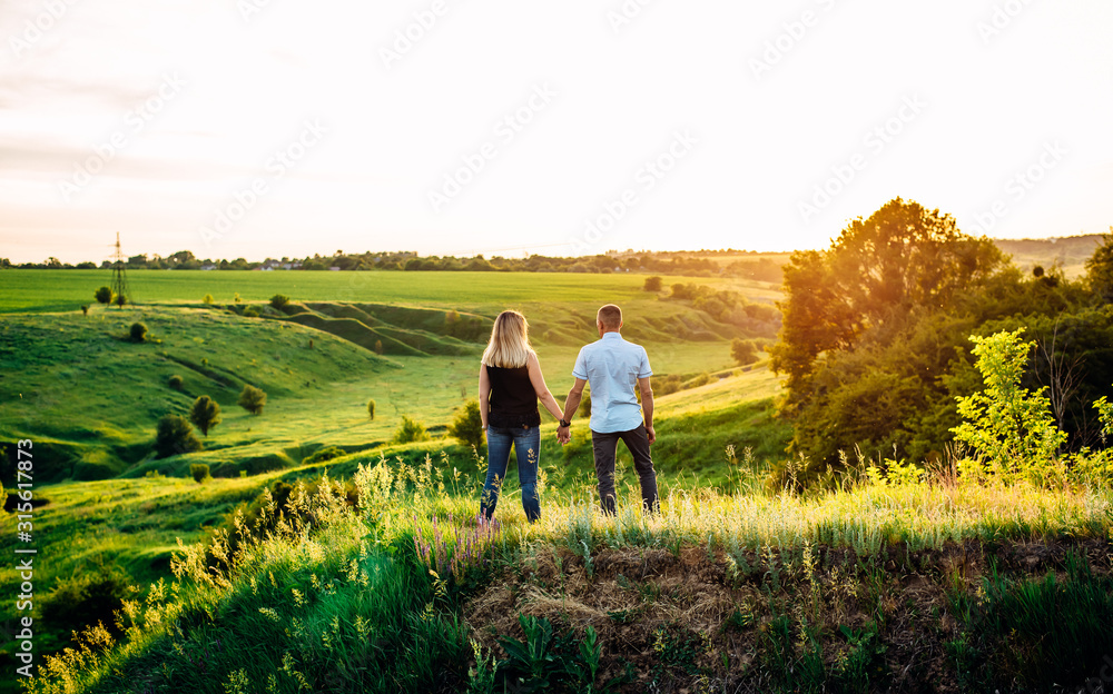 Girl and guy holding hands and standing on a green hill. Sunset. Back view.