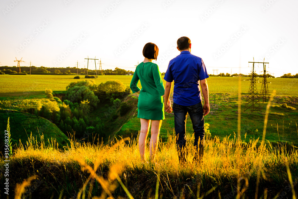 Girl and guy holding hands and standing on a green hill. Sunset. Back view.