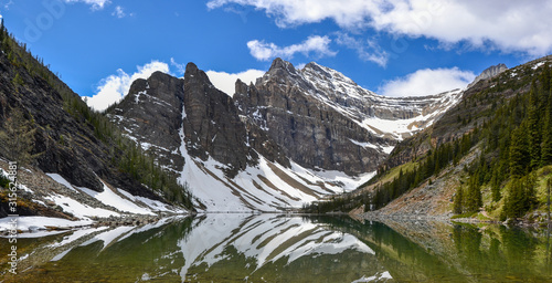 The mirror like reflections on Lake Agnes create beautiful patterns against the mountainous backdrop spotted with snow and pine trees on a sunny and partially cloudy day.