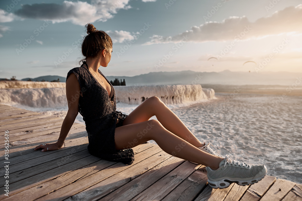 Young sexy girl in black dress sitting at natural pool Pamukkale at sunset light. Natural travertine pools and terraces in Pamukkale, Turkey