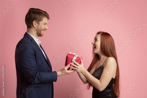 Giving a gift. Valentine's day celebration, happy caucasian couple isolated on coral studio background. Concept of human emotions, facial expression, love, relations, romantic holidays.